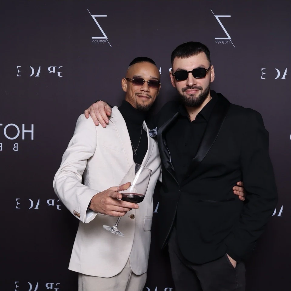 Two guests in a white suit and a black tuxedo posing together at a Hotel Zoo Berlin step-and-repeat