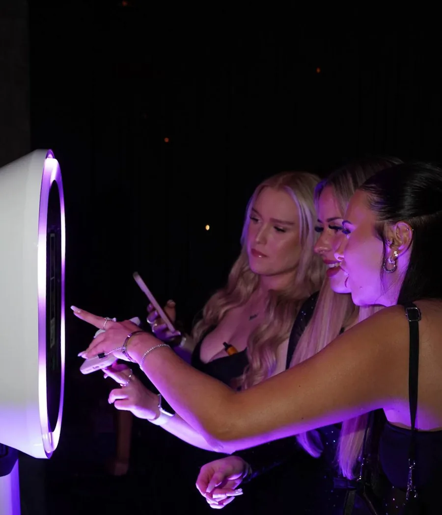 Three guests in evening wear using a purple-lit photo booth, one pointing at the screen