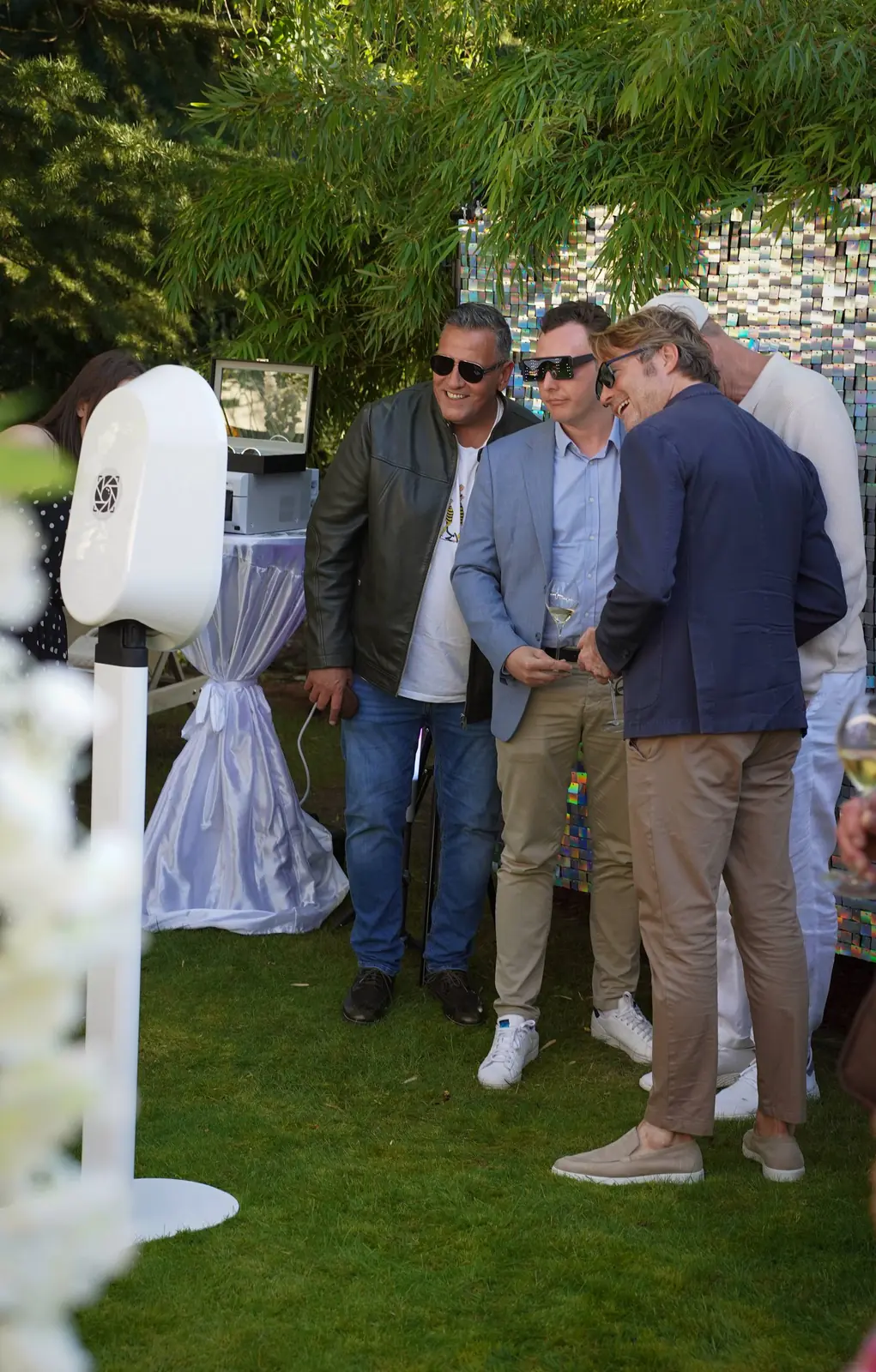 Guests laughing beside a white pedestal booth and silver sequin backdrop at a garden party