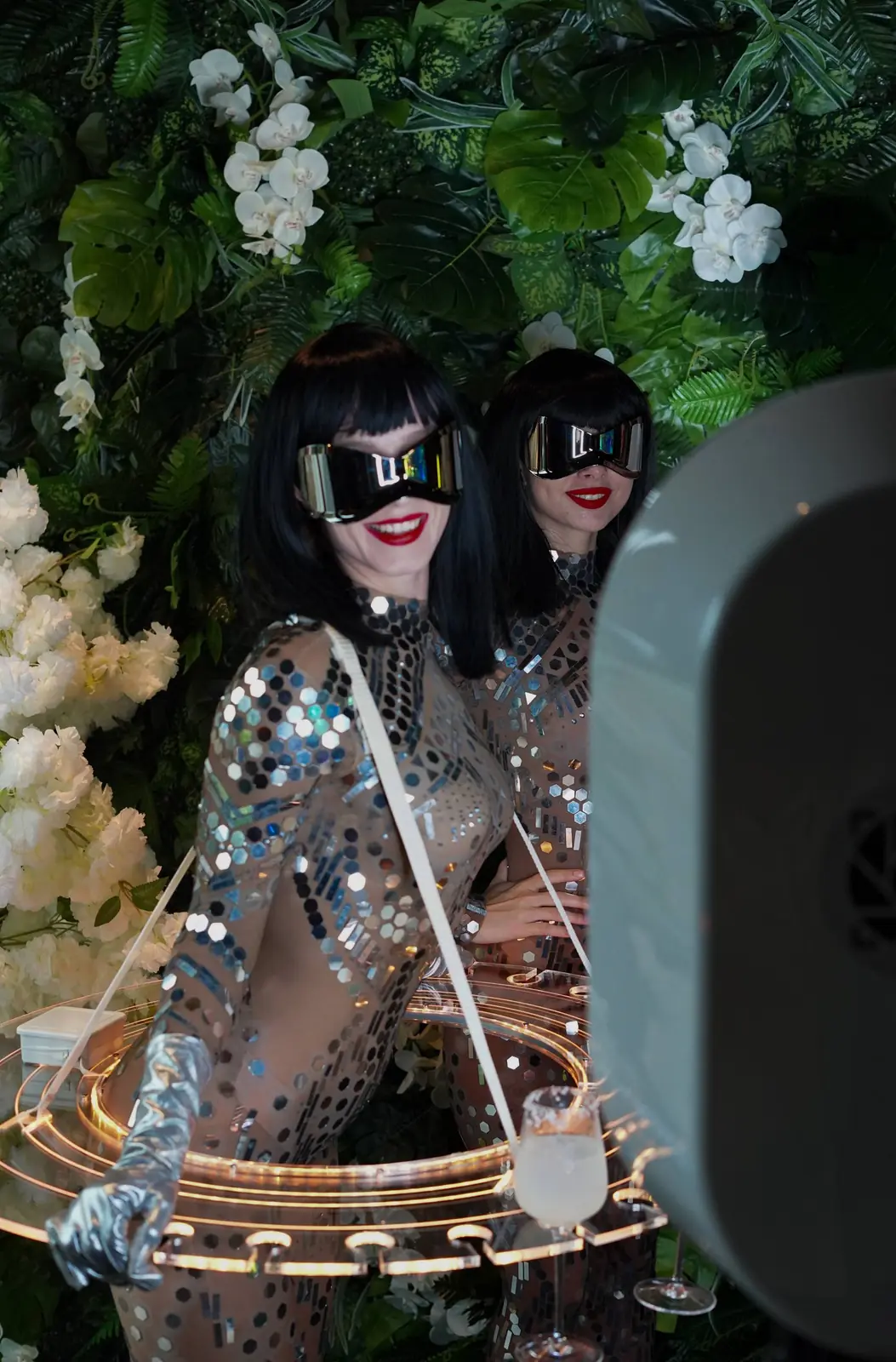 Two performers in mirrored bodysuits posing beside a photo booth and a flower wall